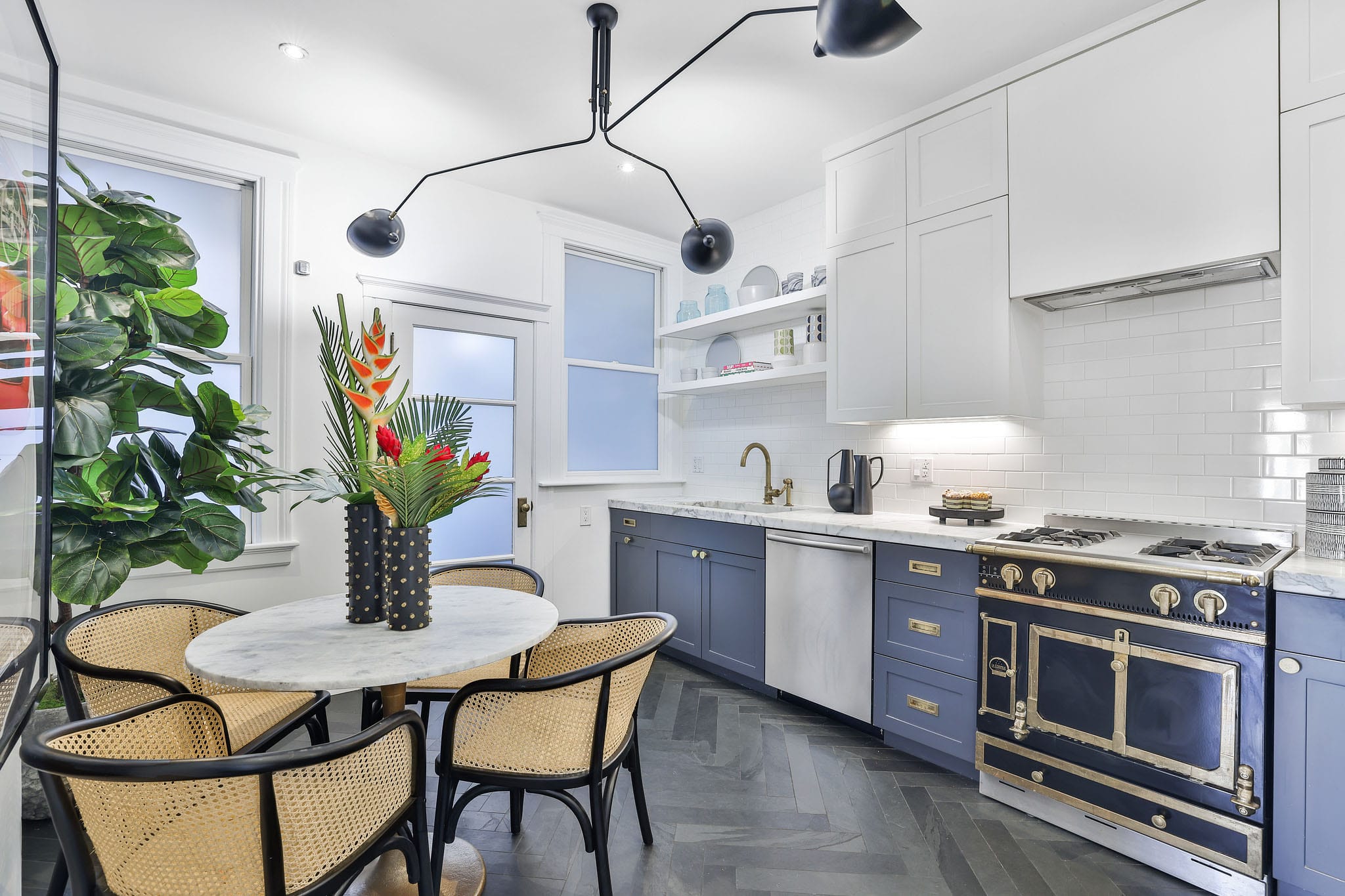 An angled view showing the dining table with rattan chairs on the left, and the dusty blue lower cabinets and white upper cabinets, alongside the black oven, on the right.