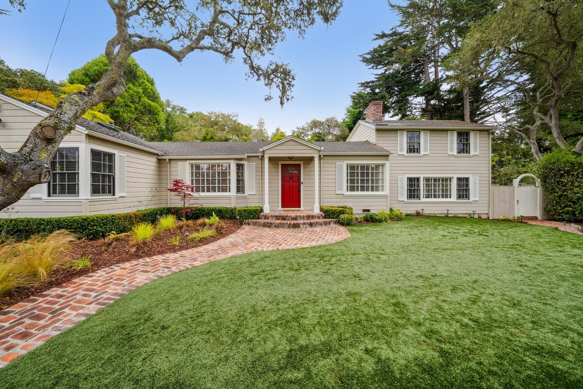 Traditional home with beige lap siding, red front door, brick path, manicured hedges, and mature tree in Carmel Woods.
