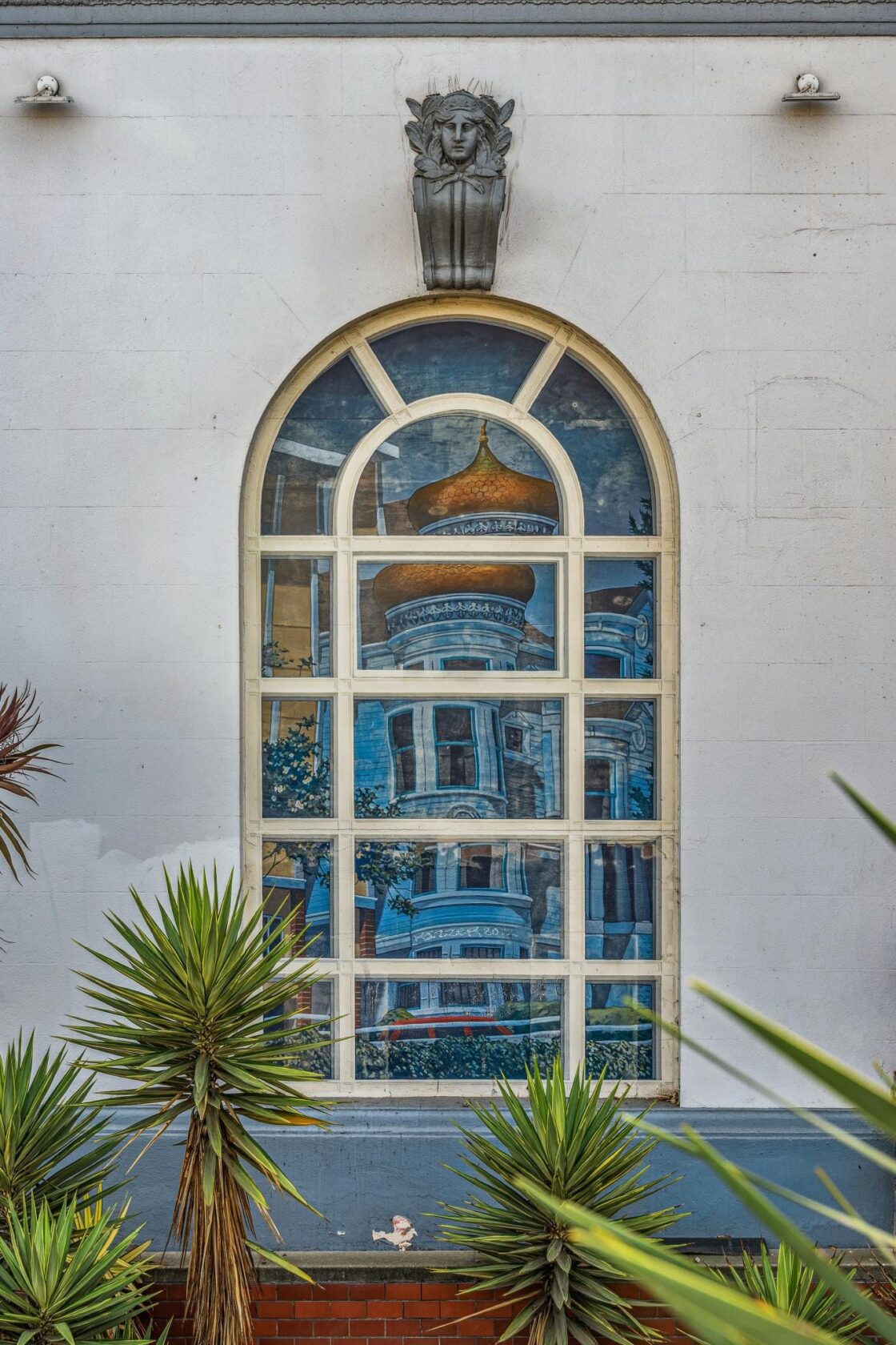 The gold onion dome and Victorian facade of the McCormick House reflected in a large arched storefront window across the street, with tropical plants in the foreground.