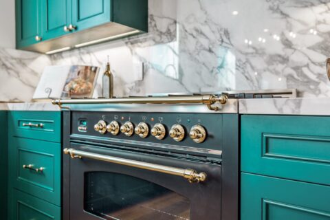 Italian Ilve black range with brass knobs and handles set against white marble backsplash and teal sapphire cabinets