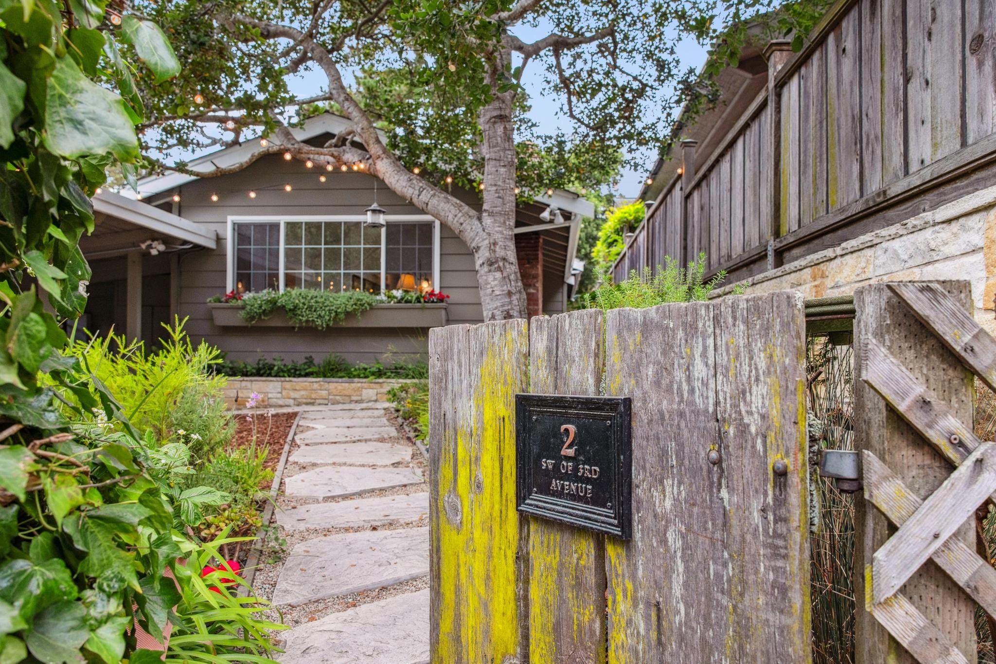 A mossy, weathered garden gate with address plaque and stone path leading to a grey cottage in Carmel-by-the-Sea.