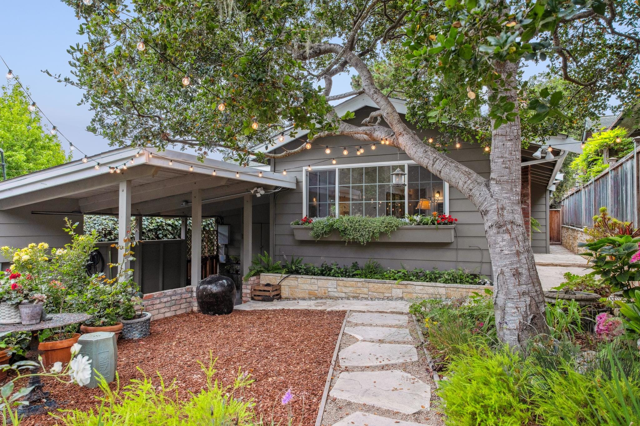 Grey cottage exterior with carport, string lights, and stone garden path in Carmel-by-the-Sea.