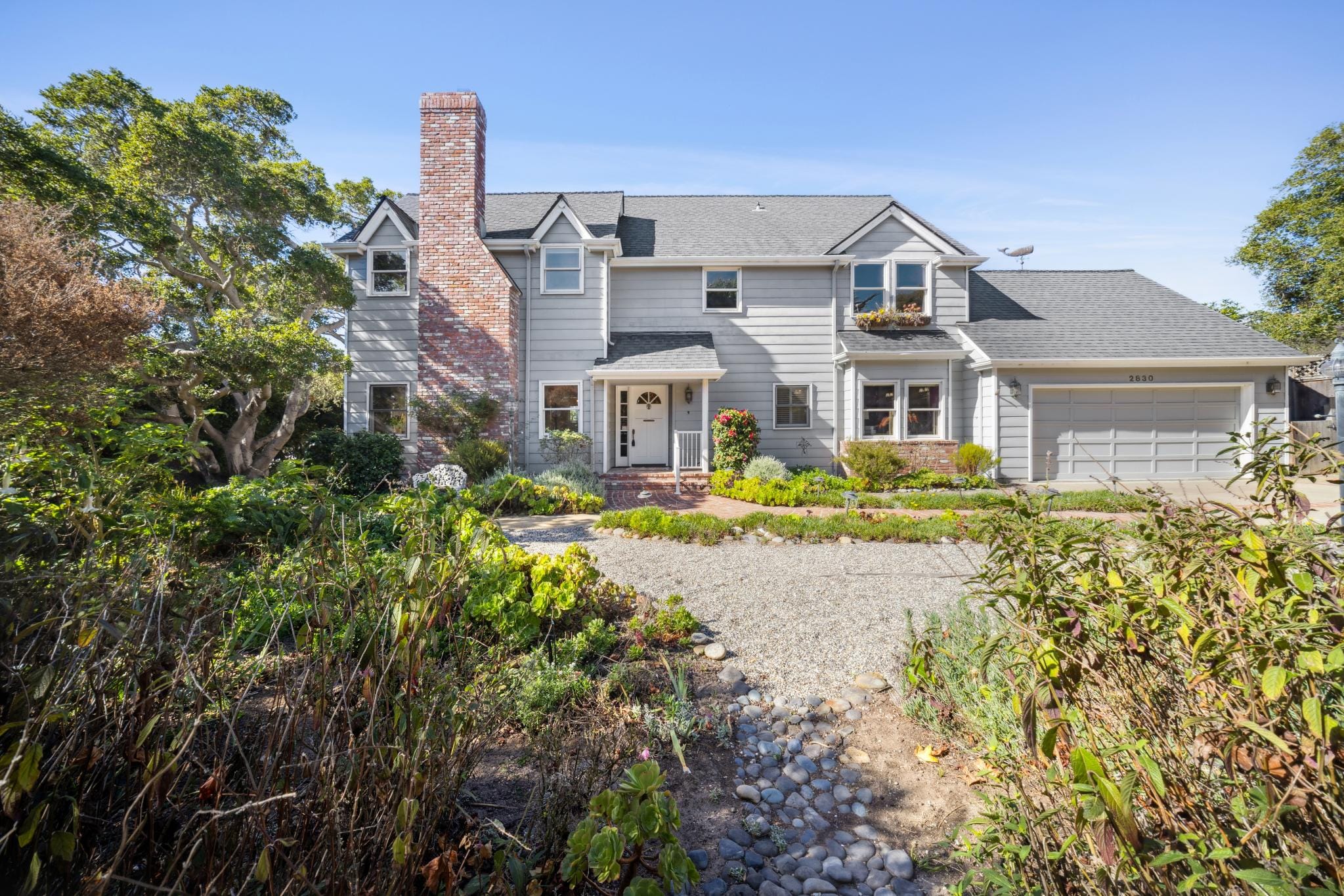 Two-story Cape Cod home with gray siding, brick chimney, gravel driveway, and lush front garden in Carmel-by-the-Sea.