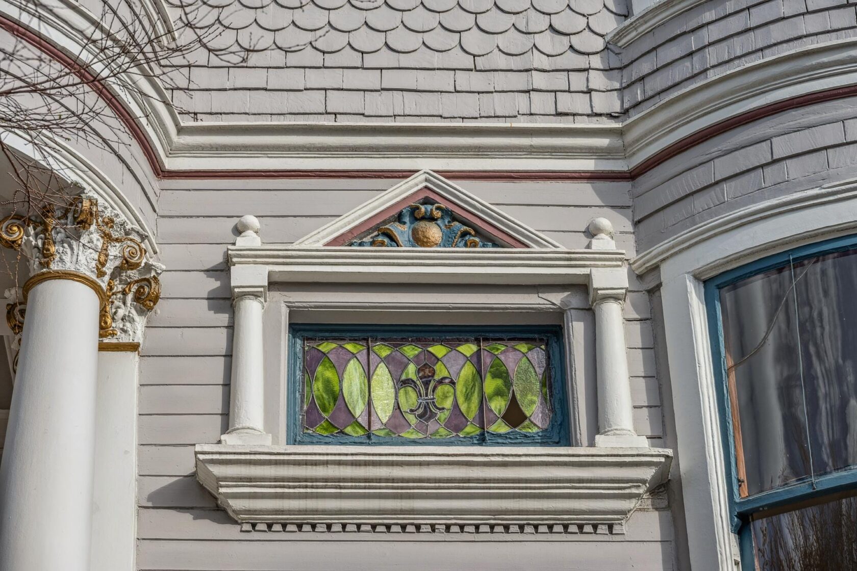 Detailed view of an original stained glass transom window set within a classical pediment, flanked by miniature columns, with a gilded Corinthian capital visible to the left.