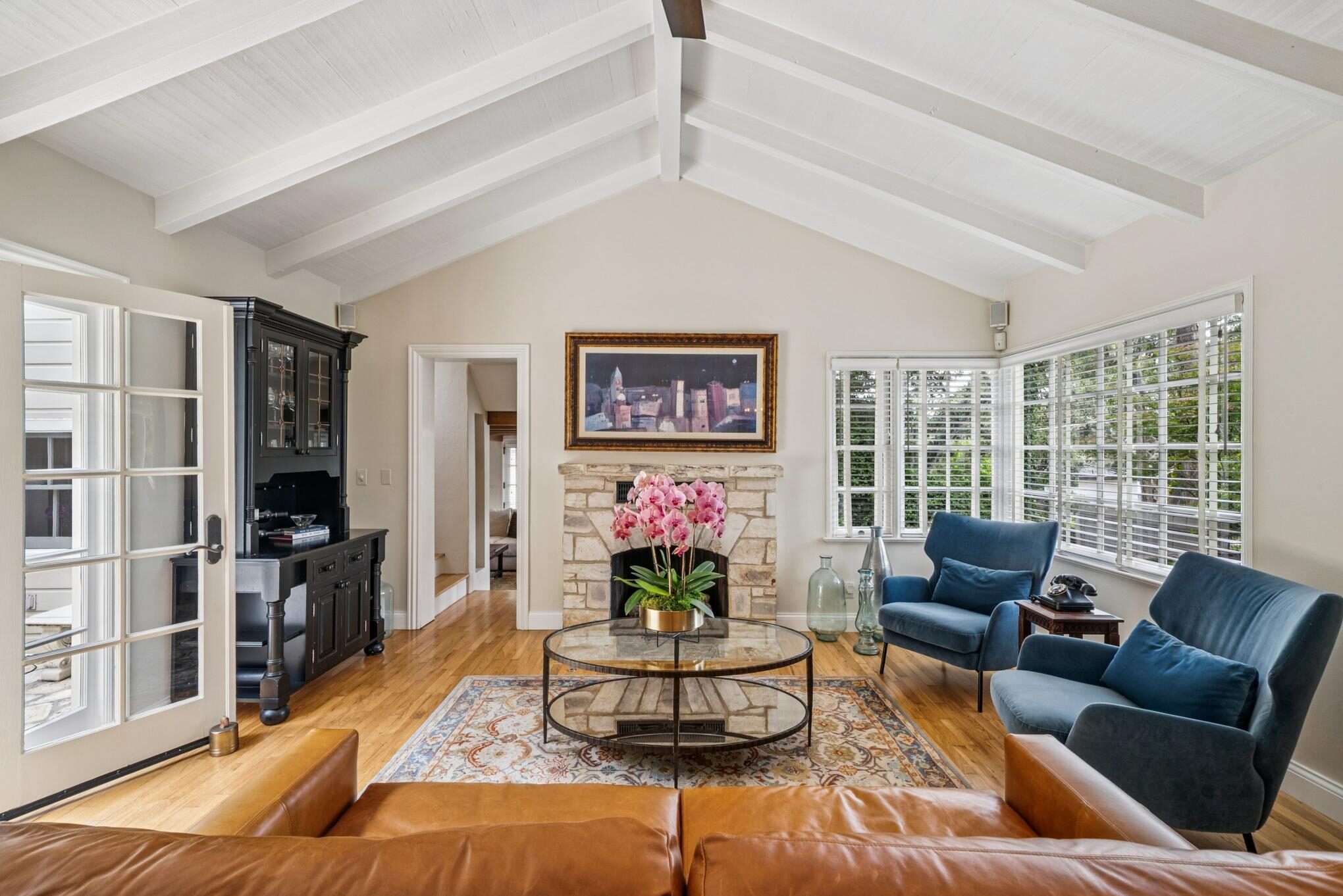 Living room with stone fireplace, vaulted beamed ceiling, hardwood floors, and French doors to patio in a Carmel Woods home.