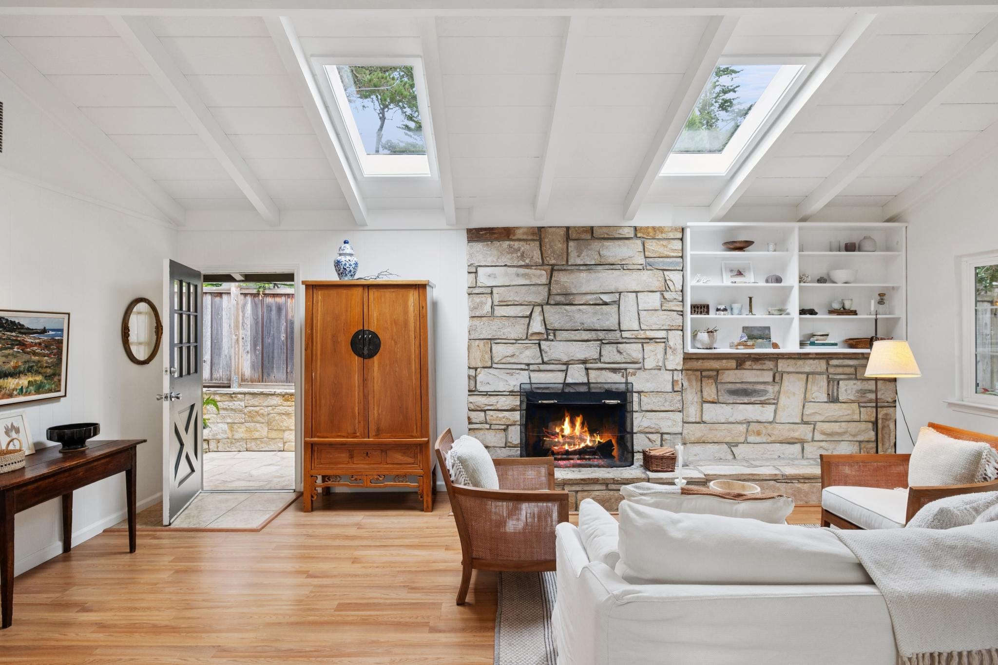 Living room with vaulted beamed ceiling, skylights, stone fireplace, and an open front door in a Carmel-by-the-Sea cottage.