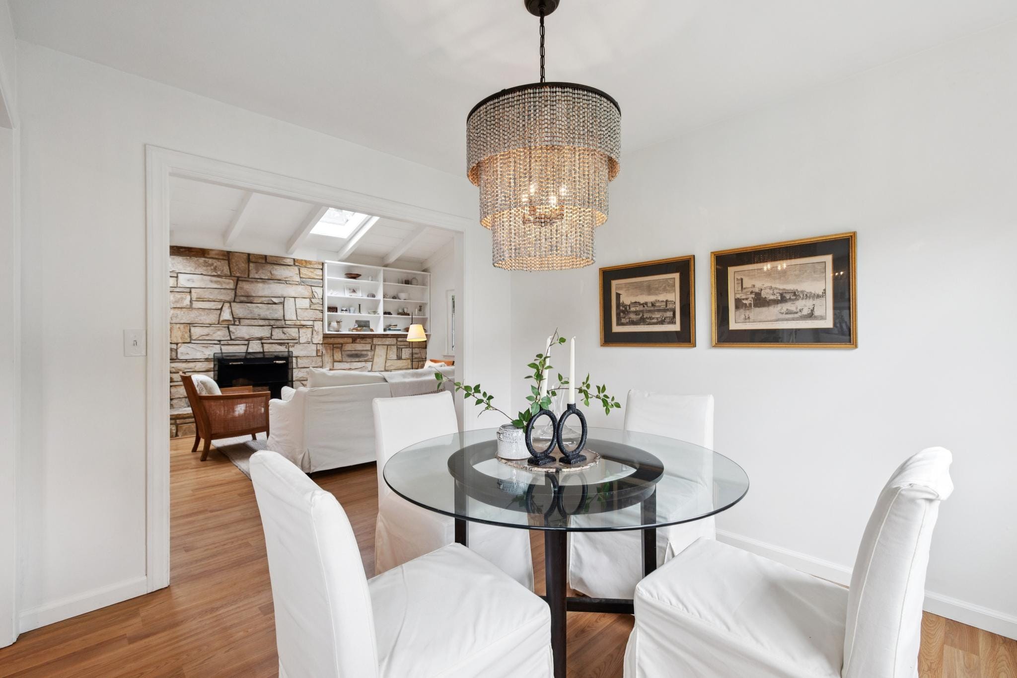 Dining room with glass table, beaded chandelier, and views into a living room with a stone fireplace in a Carmel-by-the-Sea cottage.