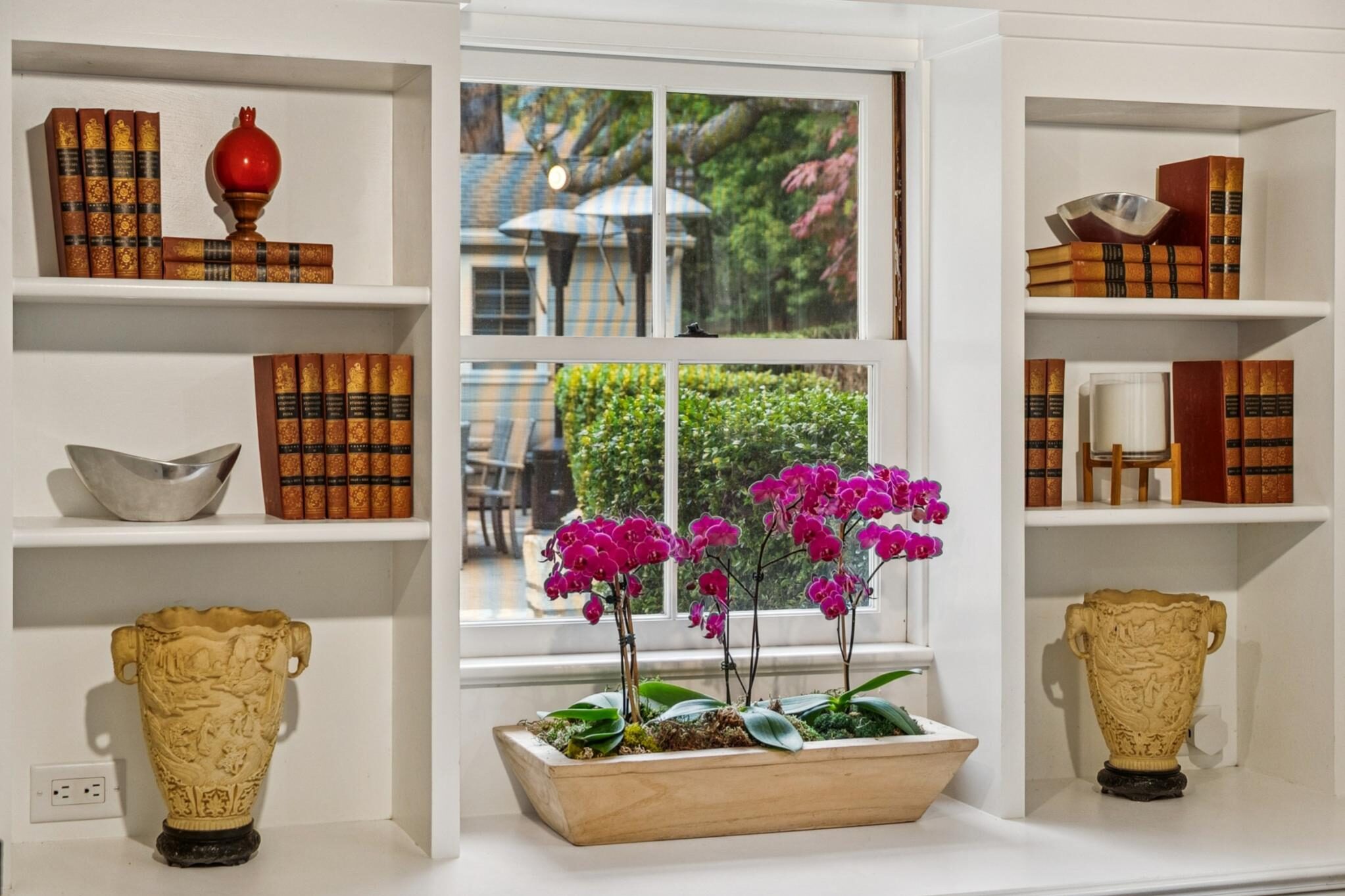 White built-in shelves with books, ceramics, and pink orchids on a windowsill overlooking a garden patio in a Carmel Woods home.