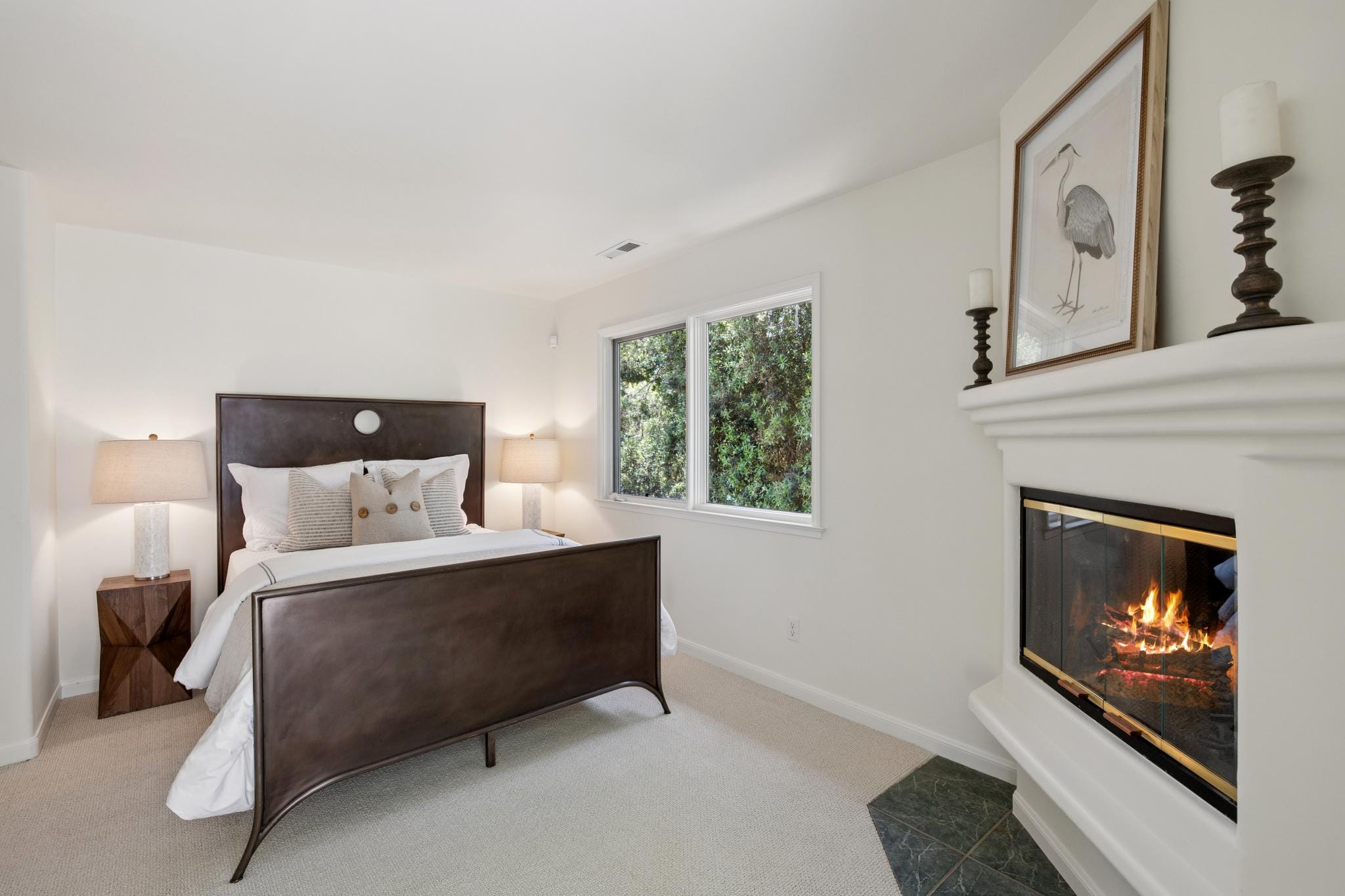 Bedroom with dark wood bed frame, fireplace with gold-trimmed doors, and garden window views in a Carmel, CA home.
