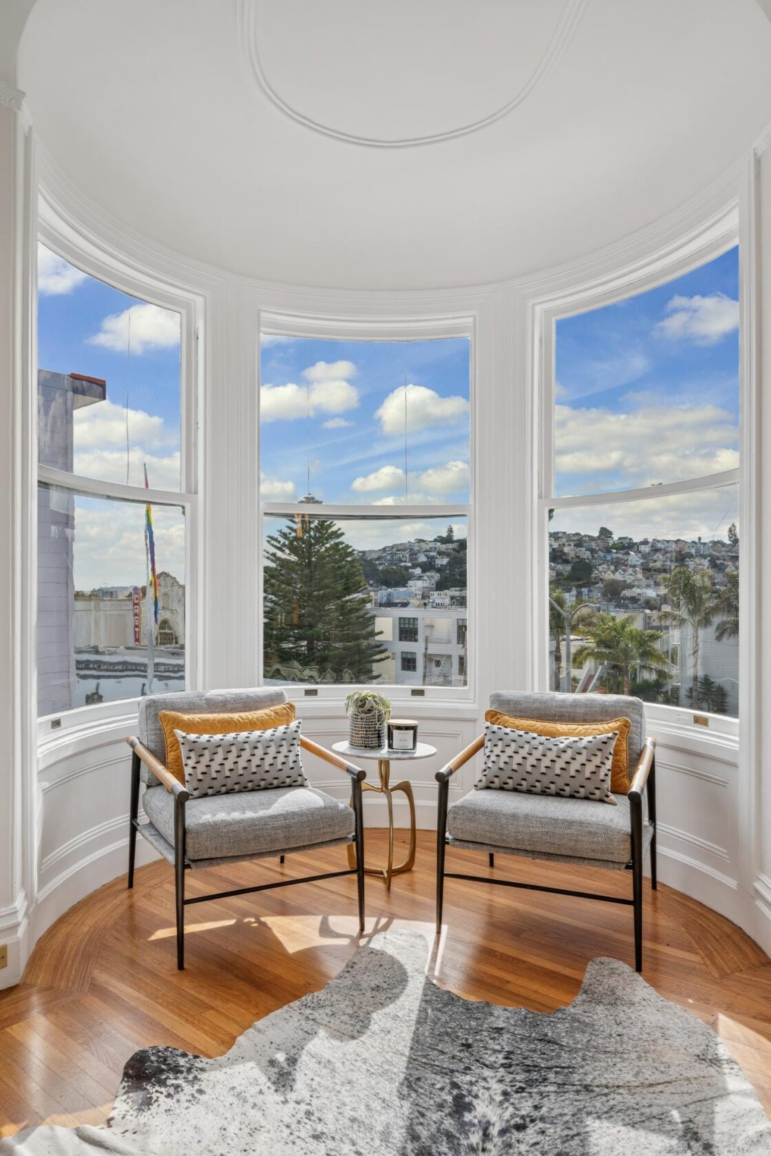 Interior of the McCormick House's curved turret bay, with wraparound wood-framed windows overlooking the Castro neighborhood, hardwood floors, and a set of chairs with a small table in the middle.