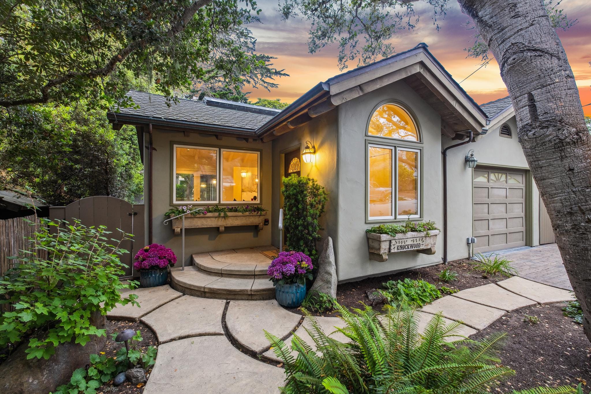 Cottage named Knockwood with arched window, lantern entry, stone path, and lush garden in Carmel, CA.