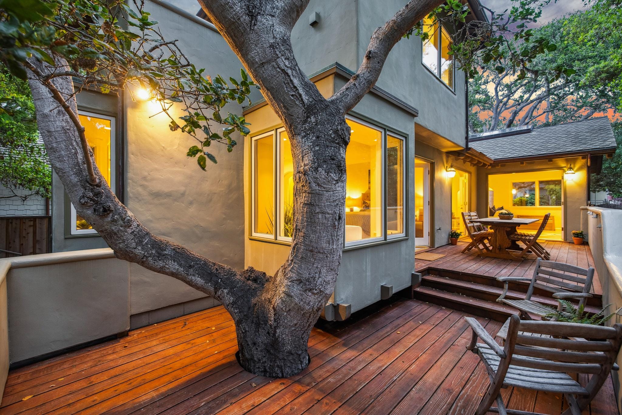 Wood deck with mature tree, outdoor dining set, and glowing interior views at a Carmel cottage at dusk.