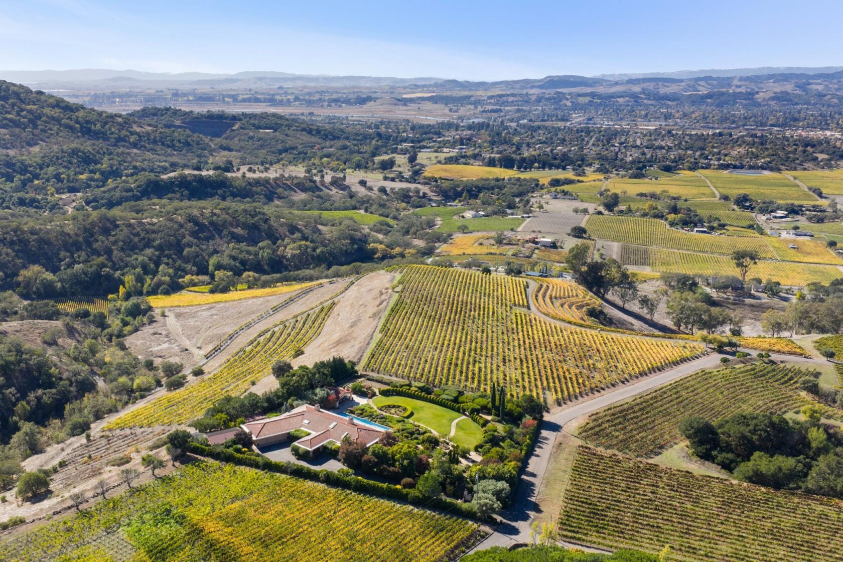 Aerial photograph of 215 Kreuzer Lane in Napa, showing a hillside estate surrounded by fall-color vineyards, with a pool, manicured grounds, and the Mayacamas Mountains visible in the background.