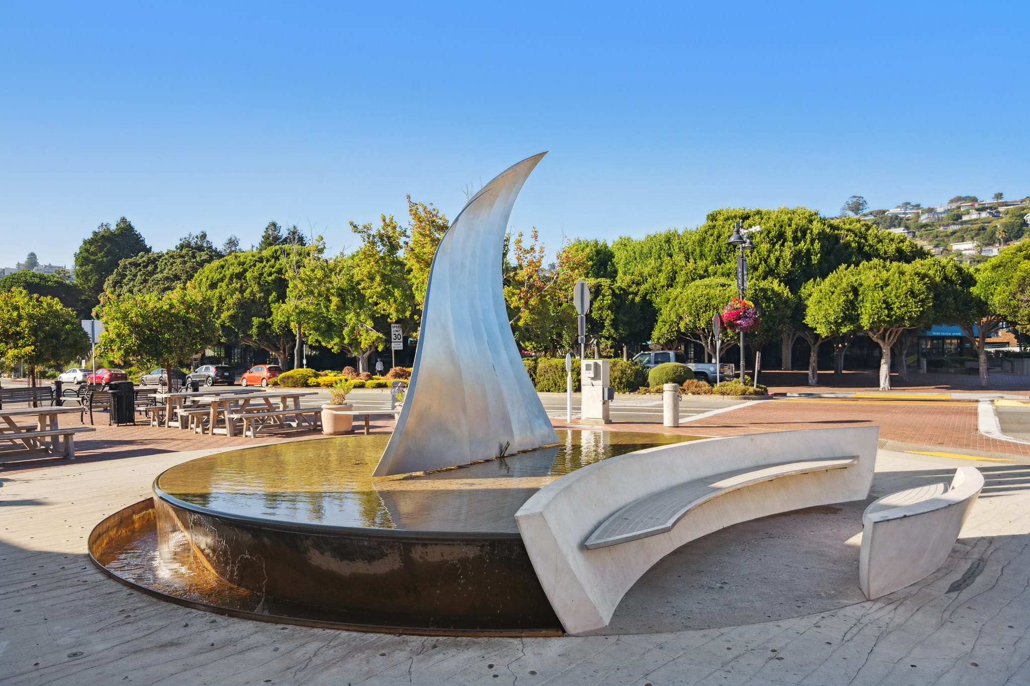 Stainless steel sailboat sculpture fountain at Tiburon Town Square with tree-lined streets and hillside homes in the background