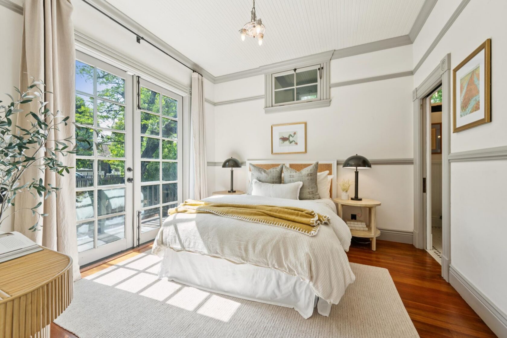 Staged bedroom with French doors, open curtains, and clear surfaces let in natural light for a listing photo