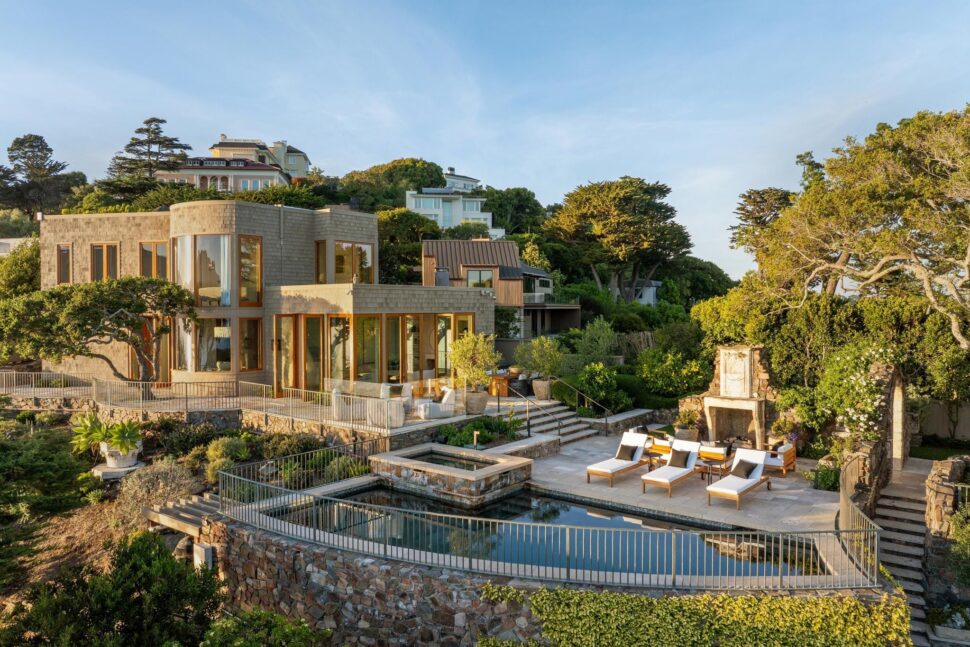 Aerial photograph of a curved stone and shingle luxury home in Belvedere with a swimming pool, spa, outdoor stone fireplace, and terraced gardens visible during golden hour.