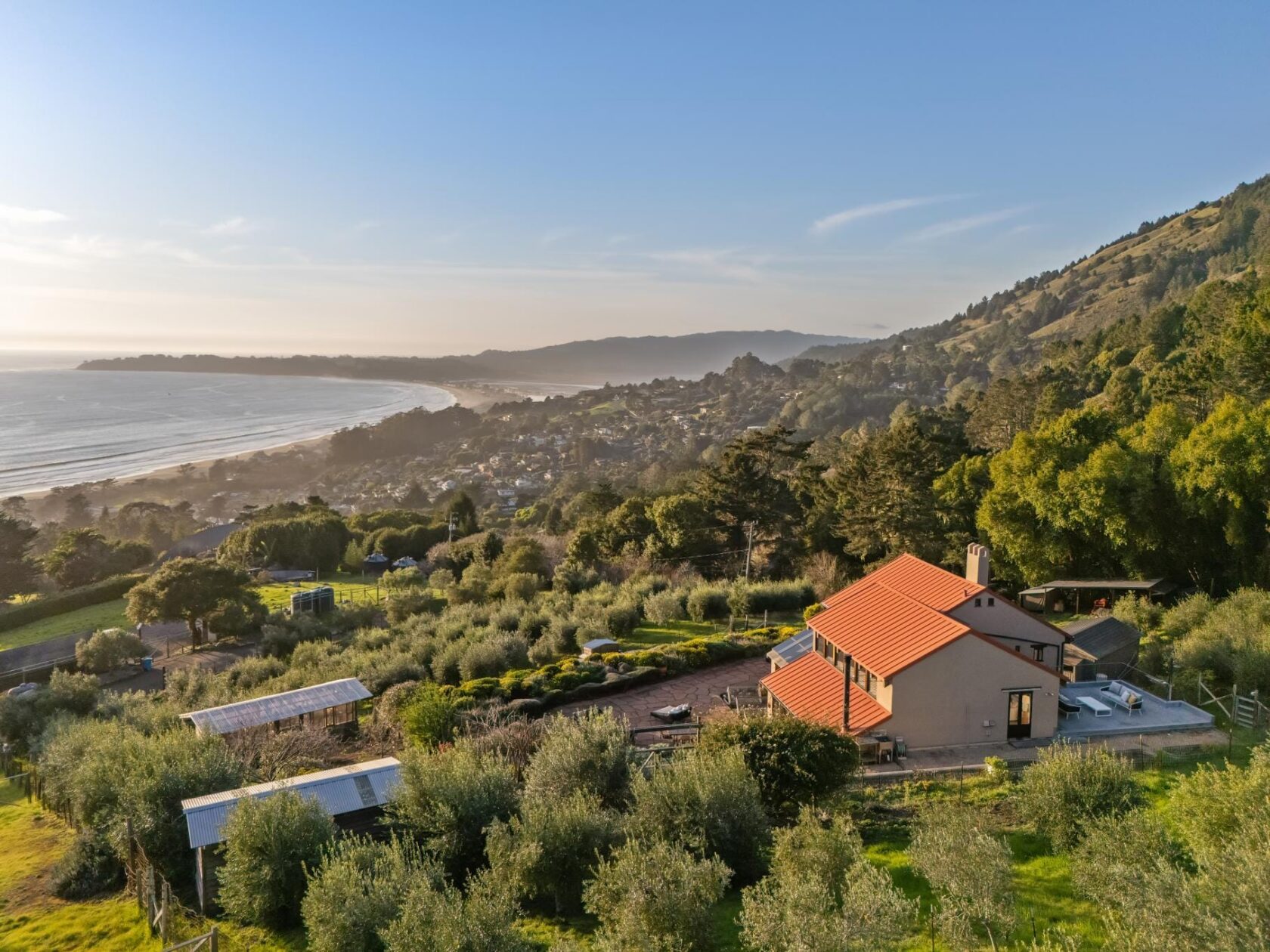 Golden hour aerial photograph of a hillside home at 6986 Panoramic Highway in Stinson Beach, with an olive orchard, outbuildings, and the Pacific Ocean and Stinson Beach curving out below.