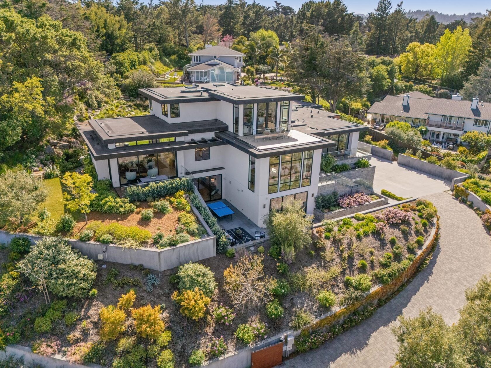 Aerial photograph of 855 Darrell Road in Hillsborough, showing a three-level contemporary estate with floor-to-ceiling windows, terraced garden beds, and a curved stone driveway set into a hillside.