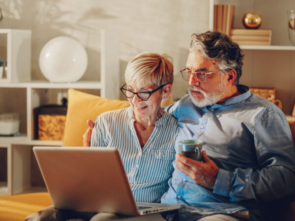 Senior married couple sitting on a couch at home and using a laptop together. Smiling mature husband and his wife are having fun while doing home shopping online